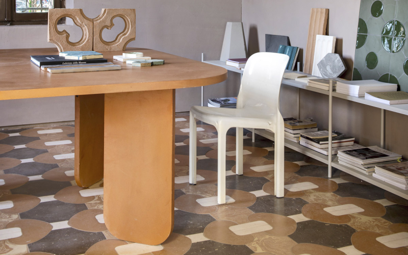 Interno di una stanza con un tavolo ovale in legno, una sedia bianca e scaffali con libri, su un pavimento a mosaico