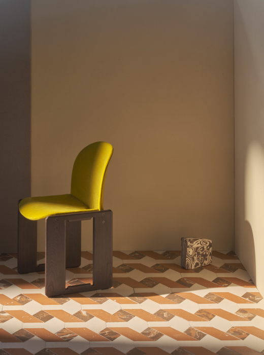 Sedia con seduta gialla su pavimento geometrico in terracotta e marmo, con un elemento decorativo sullo sfondo