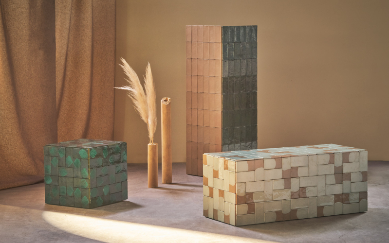 Composizione di cubi e colonne rivestiti di piastrelle colorate, con tonalità verdi e terracotta, accompagnata da rami decorativi e un sipario sullo sfondo