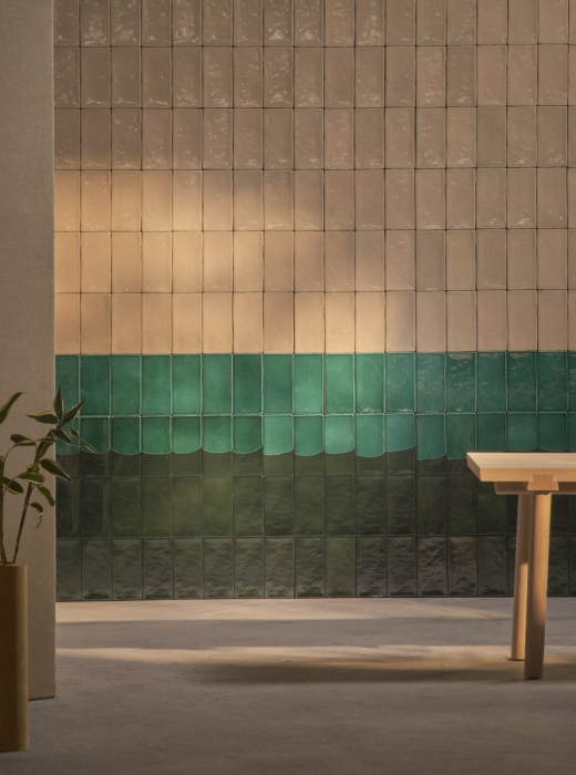 Parete decorativa con piastrelle sfumate dal beige al verde scuro, accompagnata da un vaso cilindrico con pianta e un tavolo in legno minimalista
