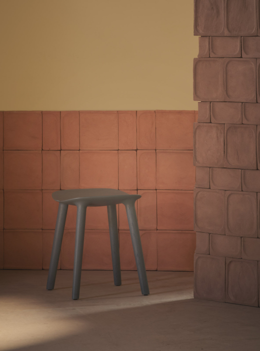 Parete in terracotta con moduli concavi, sgabello grigio e luce naturale che crea un contrasto di ombre