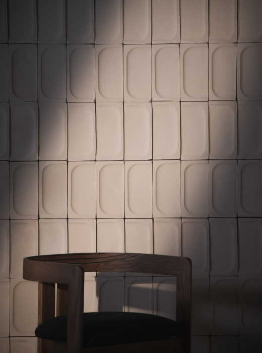 Parete in ceramica bianca con moduli concavi e una sedia in legno in primo piano, illuminata da luce naturale